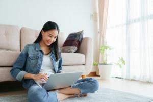 穿着牛仔裤的亚洲女孩坐在家里的地板上玩笔记本电脑. 自由职业女孩正在社交媒体上与同事进行视频会议. 概念工作在家和新常态